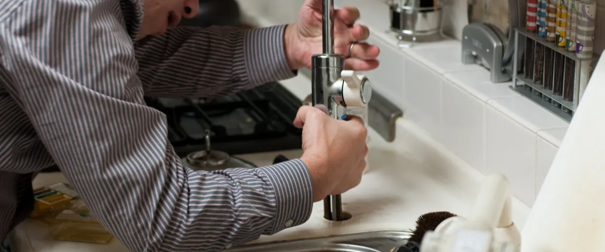 A man in a striped shirt installs a new chrome kitchen faucet into a white sink counter.