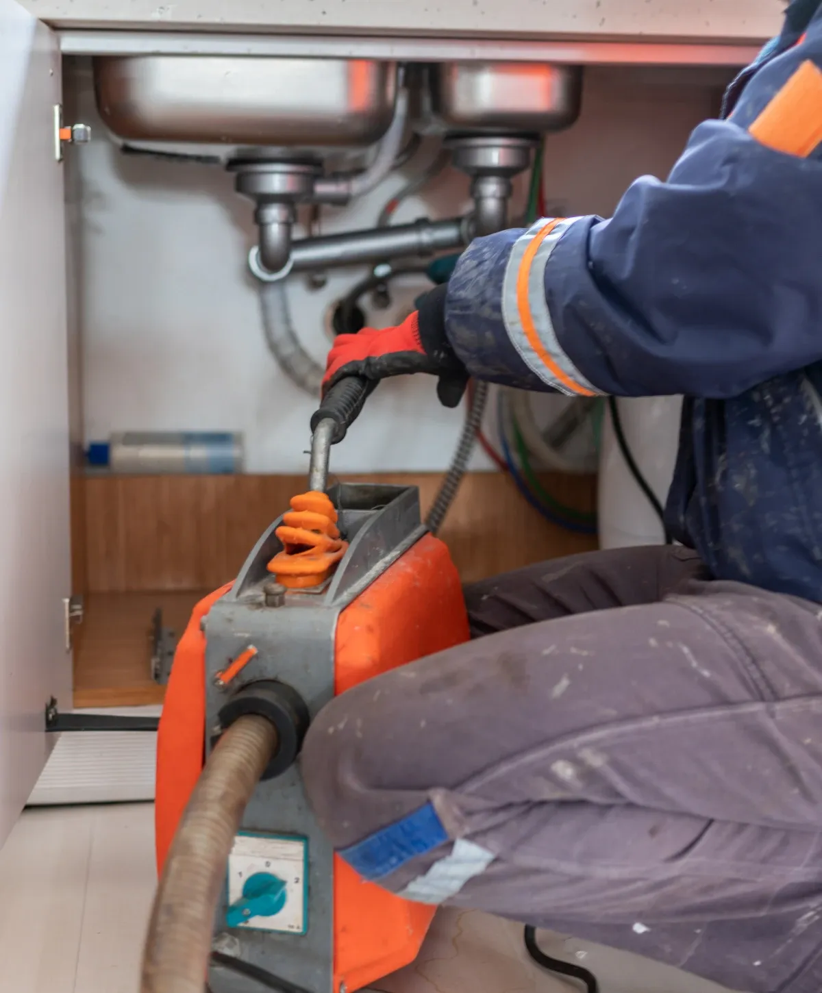 A person in work clothes and red gloves operates an orange and gray drain cleaning machine under a kitchen sink.
