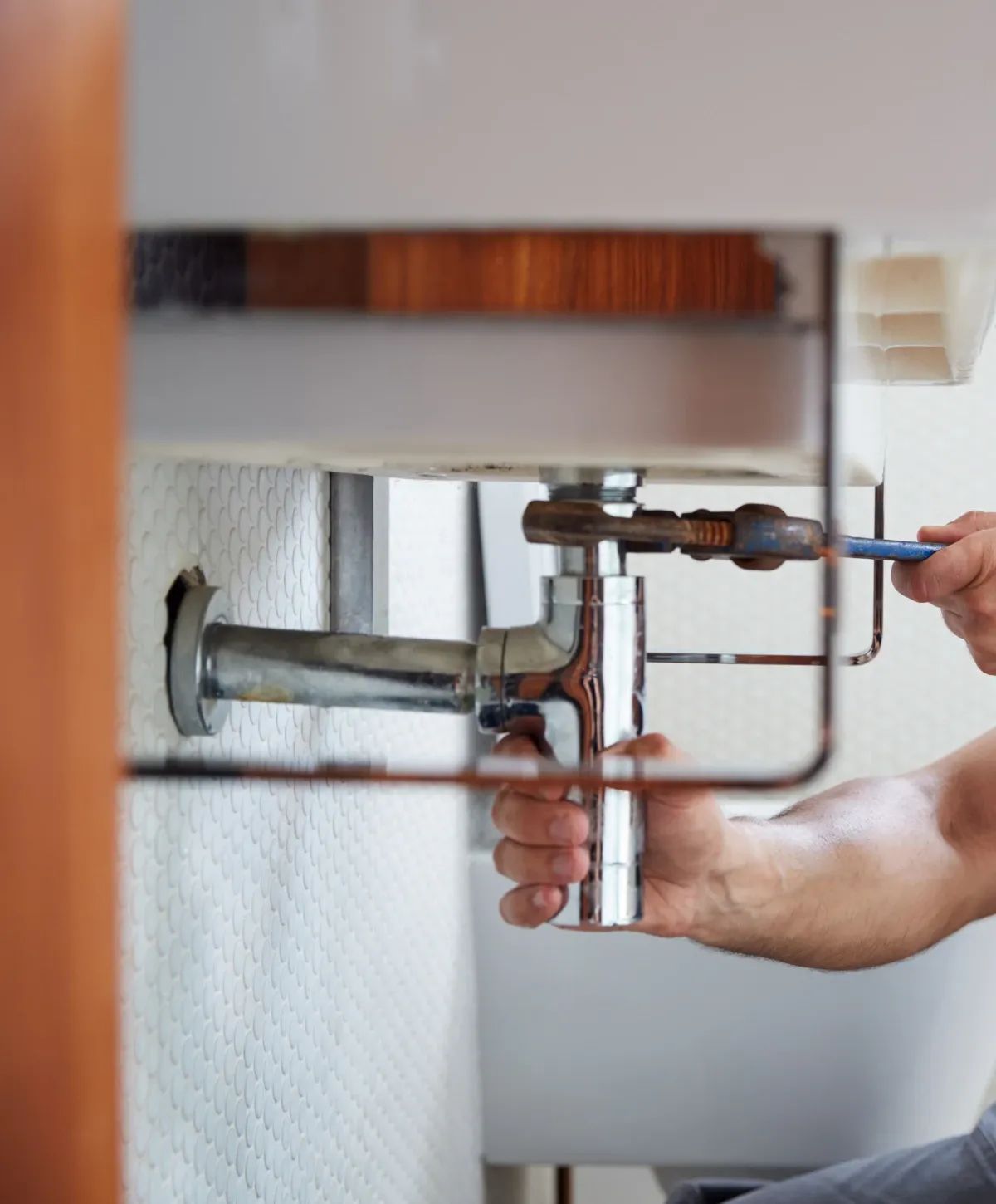 A close-up view from below shows a person's hands using a wrench to fix chrome pipes under a bathroom sink.