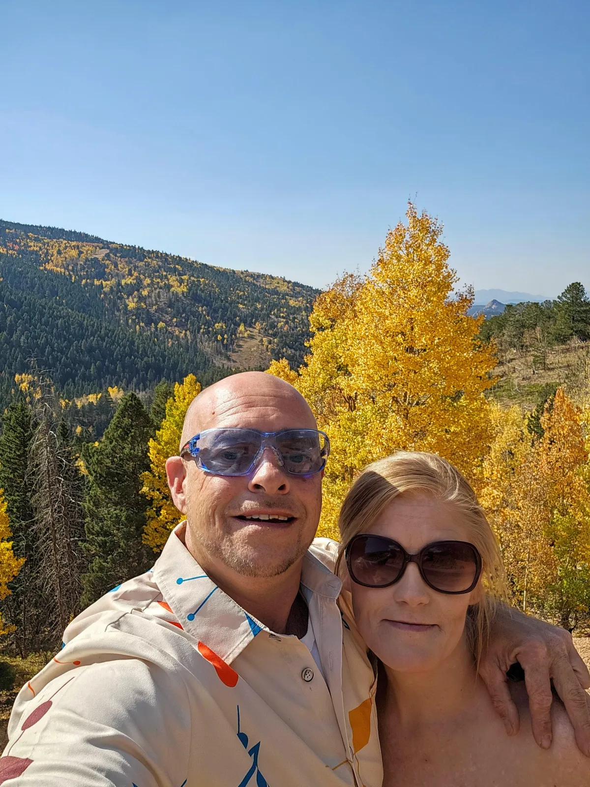 "Couple taking a selfie outdoors with a scenic mountain landscape in the background featuring autumn trees with vibrant yellow foliage under a clear blue sky."
