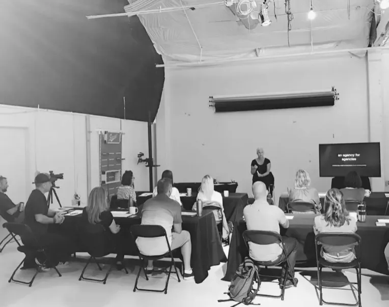 Black-and-white photo showing a group of people having a discussion around small tables in an office environment, featuring large 'world' graphics on the background wall, all framed by a pale teal square border
