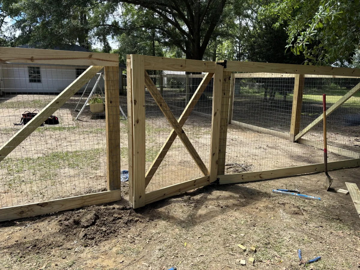 Newly installed wooden fence adjacent to a brick house, showcasing craftsmanship and attention to detail, with tools and landscaping visible in the foreground.