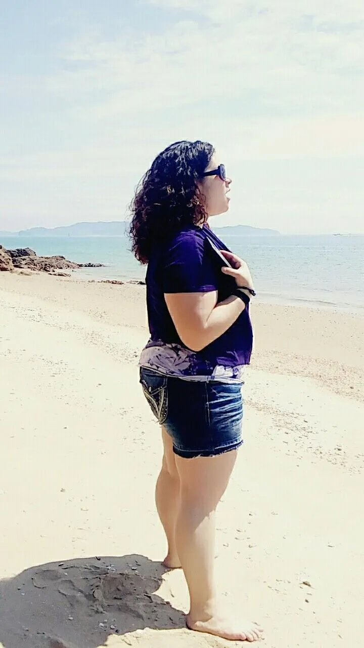 Jess DuBose standing on a sunny beach in profile, holding a phone to her chest with one hand, singing while facing the ocean, with distant mountains visible across the water