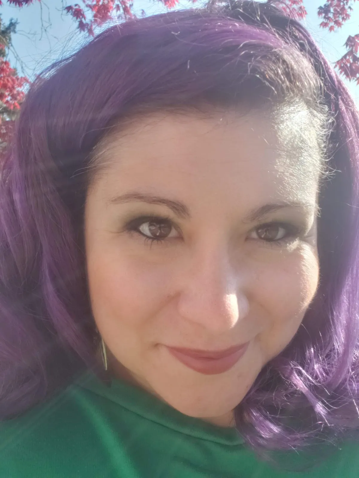 Jess DuBose smiles softly at the camera under bright sunlight, with purple hair and a natural background of red leaves and a soft blue sky. Light glows on her face and hair.