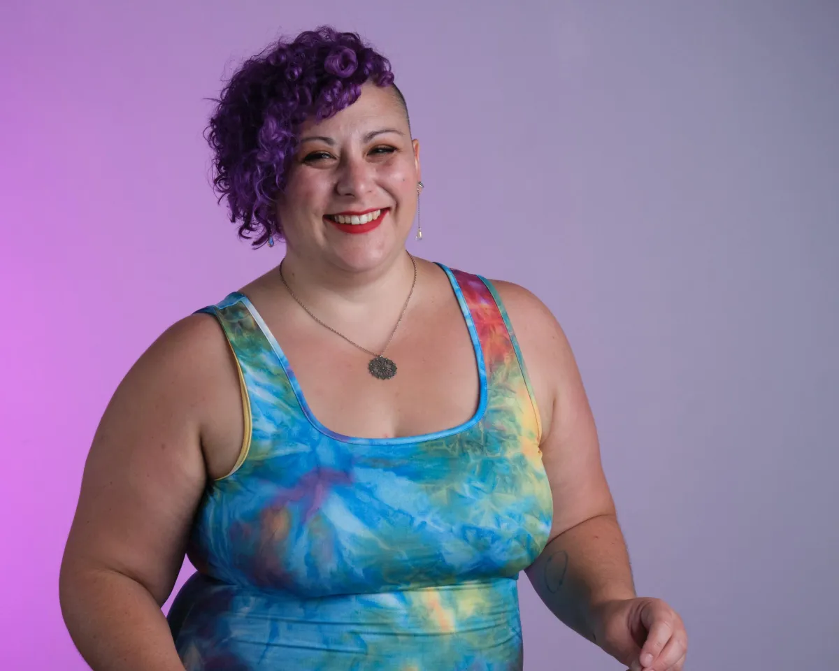 Young woman with colorful hair clips and space buns resting her head on an old TV decorated with stickers, wearing a sheer star-patterned top and looking directly at the camera against a pale blue background