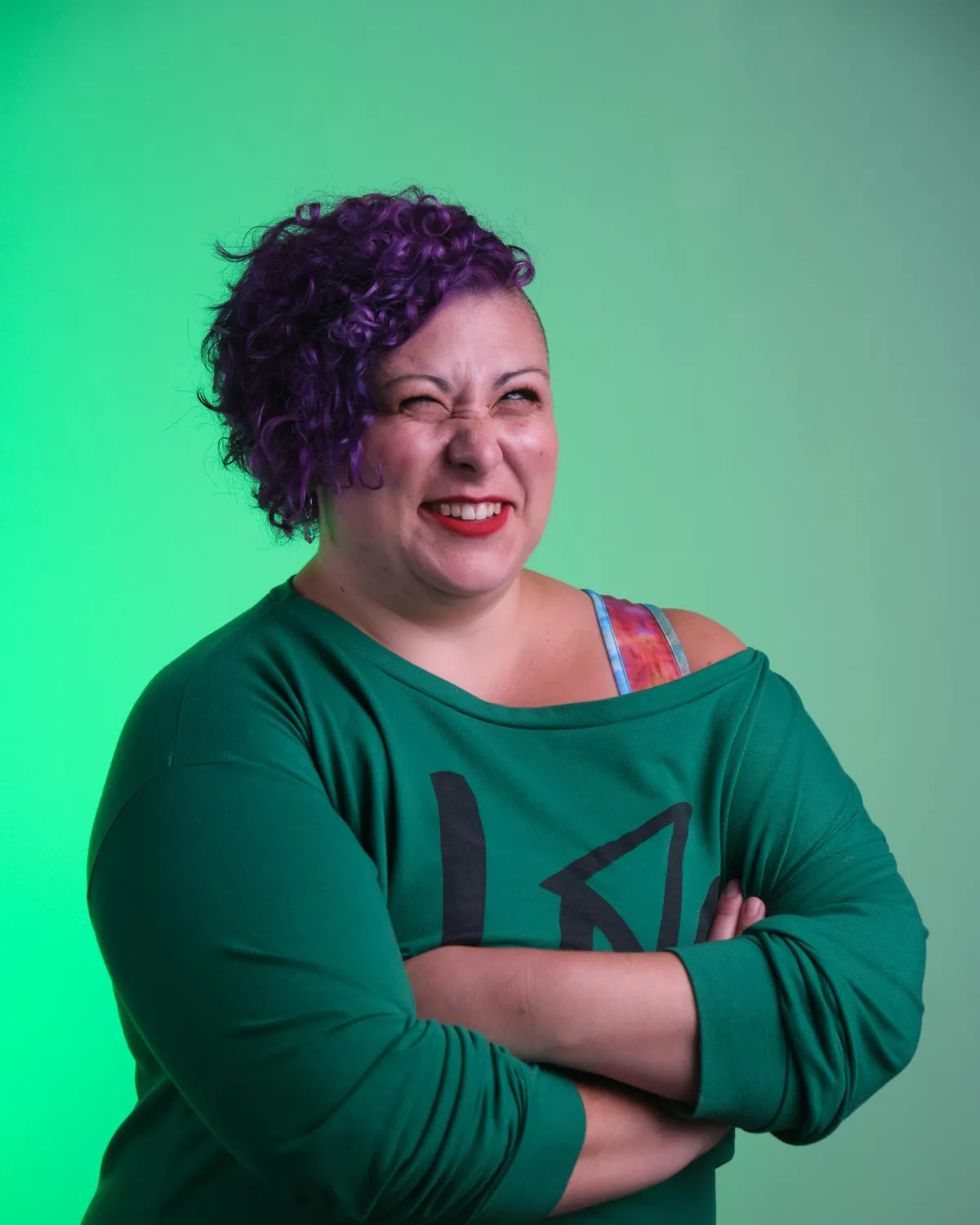 Jess DuBose stands against a green gradient background, smiling with a playful squint and arms crossed. She has bright purple curls, red lipstick, and wears a green off-shoulder sweatshirt.