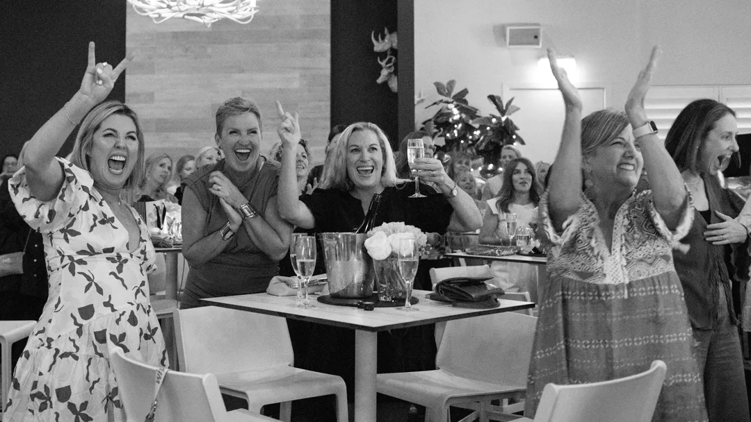 Black and white photo of three women laughing, cheering, and holding up drinks in a crowded venue