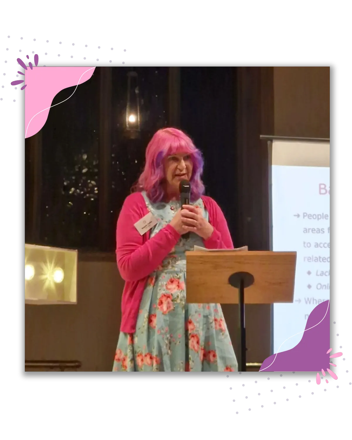 Melanie speaking on stage at School for Social Entrepreneurs. Holding a microphone, wearing a pink cardigan and has pink and purple hair.