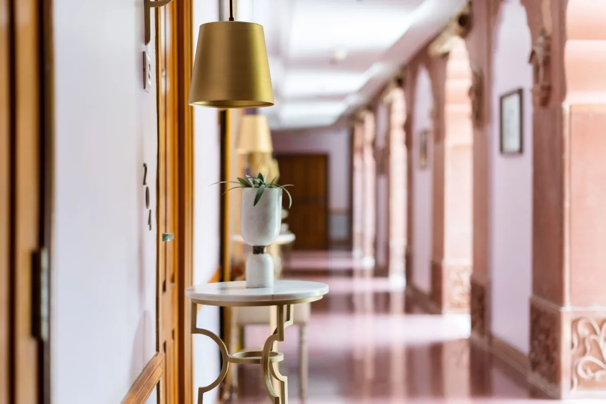 A quiet, sunlit corridor with arched doorways, a brass lamp and a small table with a plant