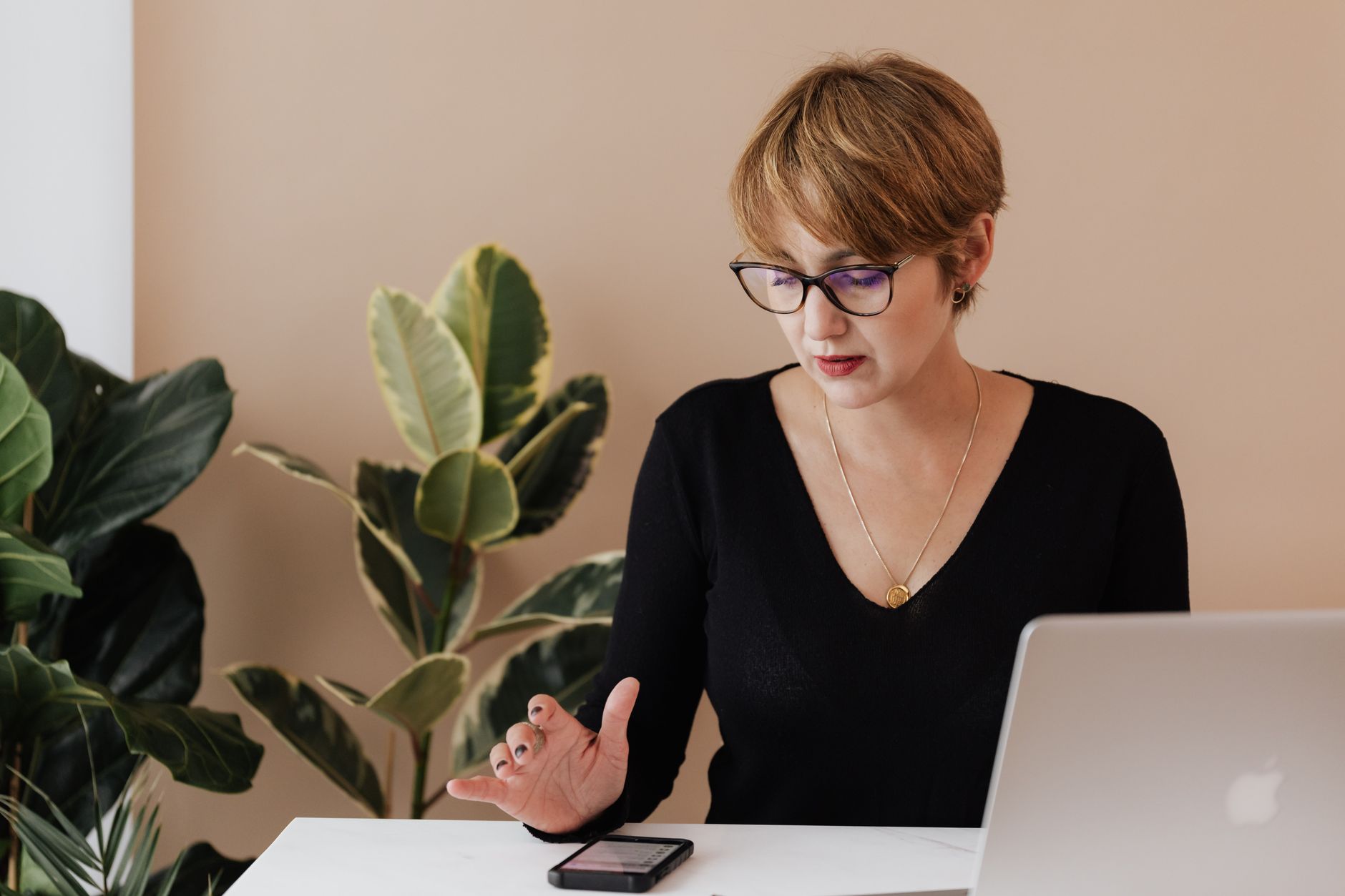 Concentrated female manager in casual outfit and eyeglasses reading message on smartphone while sitting at table with laptop in cozy workspace