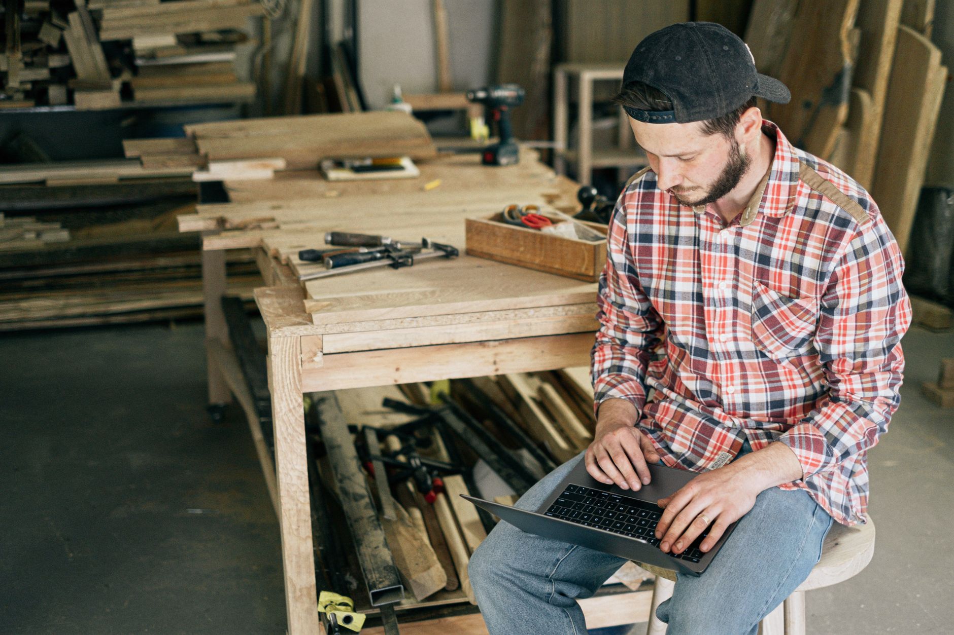 A carpenter working on his laptop in a wood workshop, surrounded by tools and materials.