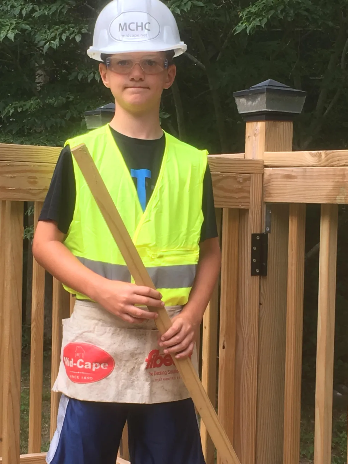 A young boy wearing a safety helmet, protective glasses, and a bright yellow safety vest stands on a wooden deck. He holds a piece of wood and wears a tool apron, engaged in a construction project