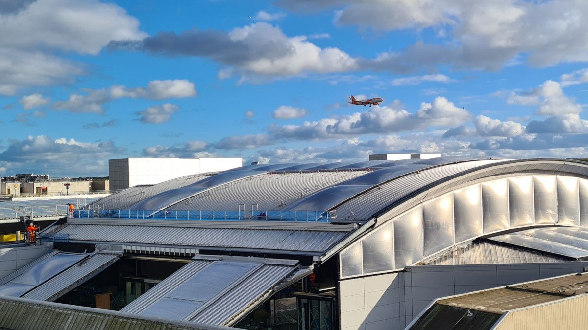 London Gatwick's terminal under a bright sky with an aircraft departing overhead — the UK arrival point for Uganda's only nonstop produce service.