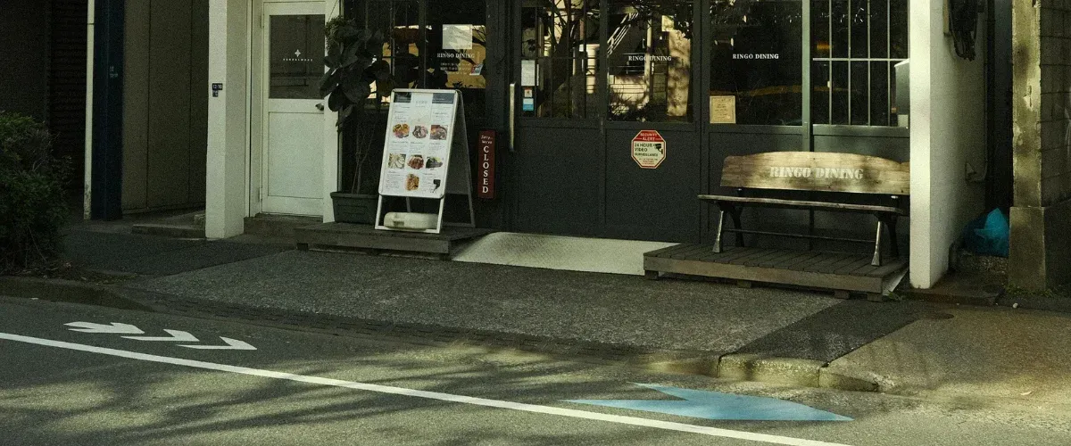 A daytime view of the Ringo Dining storefront with a menu board, wooden bench, and street arrows.