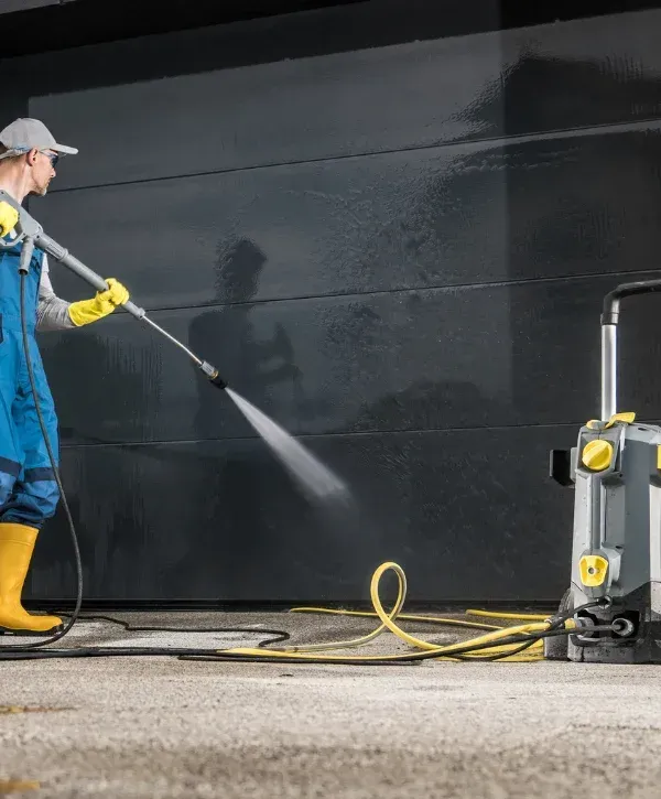 A man in blue overalls and yellow boots power washes a dark gray garage door, with a pressure washer unit nearby.
