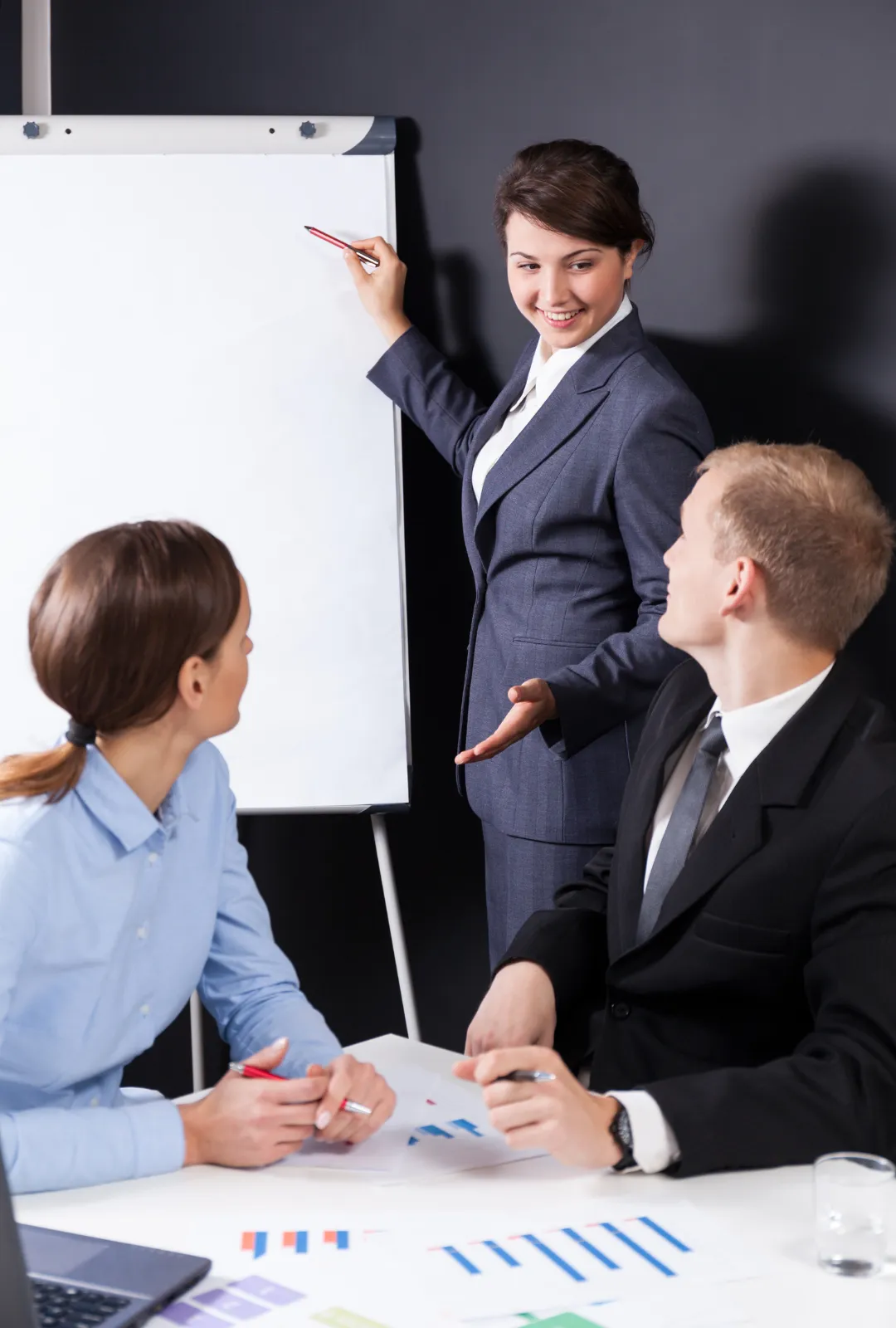 Ejecutiva señalando una pizarra mientras lidera una reunión con profesionales, representando mentoría ejecutiva y alto rendimiento empresarial