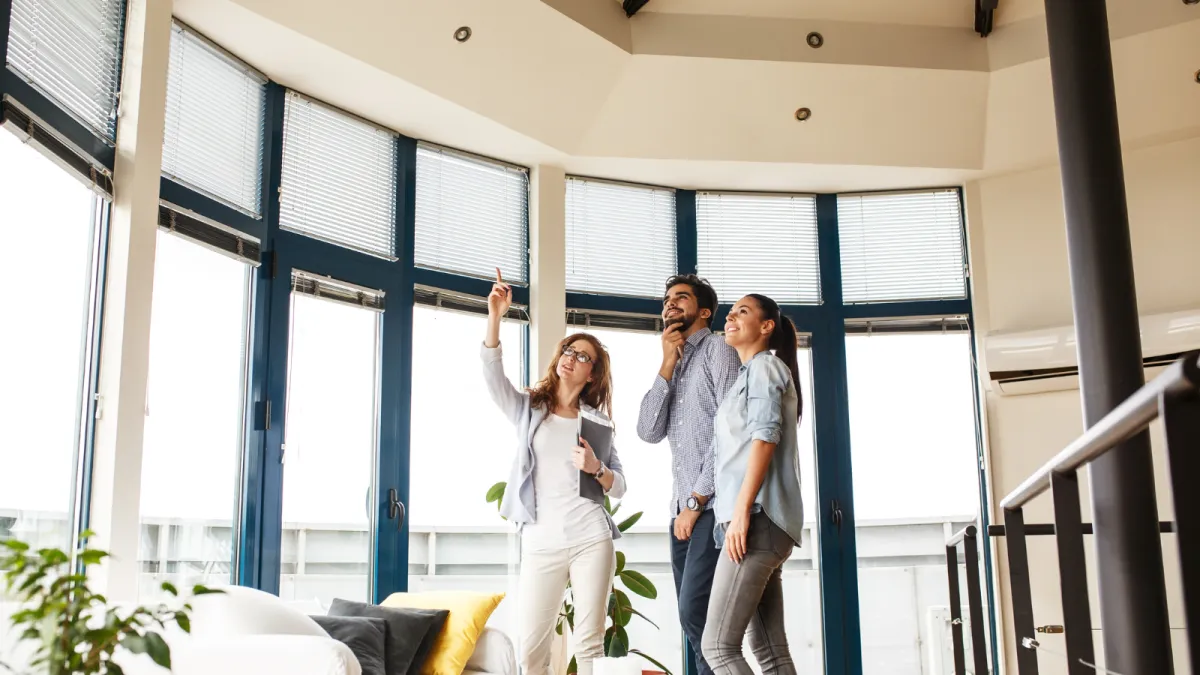 Real estate agent showing a modern home to a young couple in a bright, spacious living room with floor-to-ceiling windows