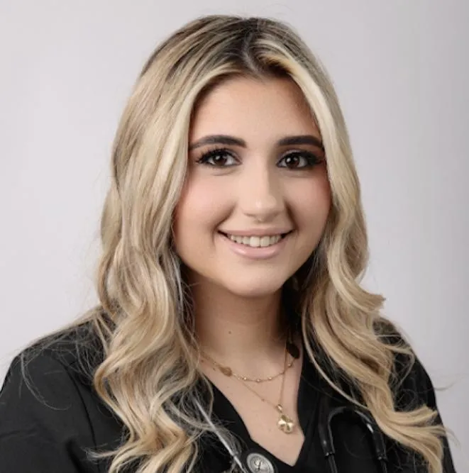 Portrait of Rena Berry, BSN, RN: a warm, professional woman in her late 30s, wearing a black medical uniform with gold trim, standing in a softly lit medspa room with neutral tones and elegant decor.