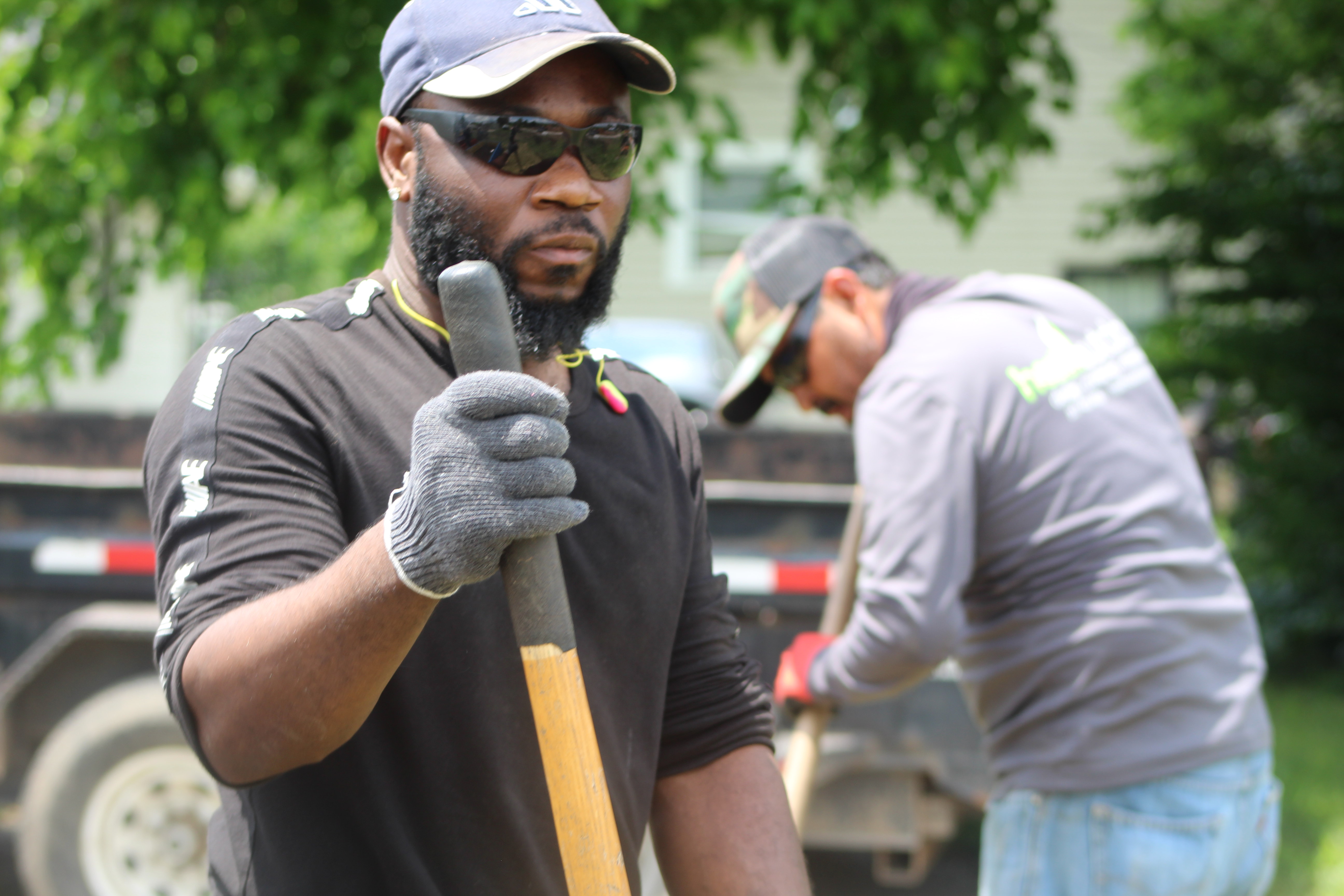 Aerial view of a diverse group of adults sketching landscape plans on large paper outdoors, surrounded by vibrant plants and garden tools, with sunlight filtering through trees. The group is engaged and smiling, representing a collaborative, hands-on learning environment.