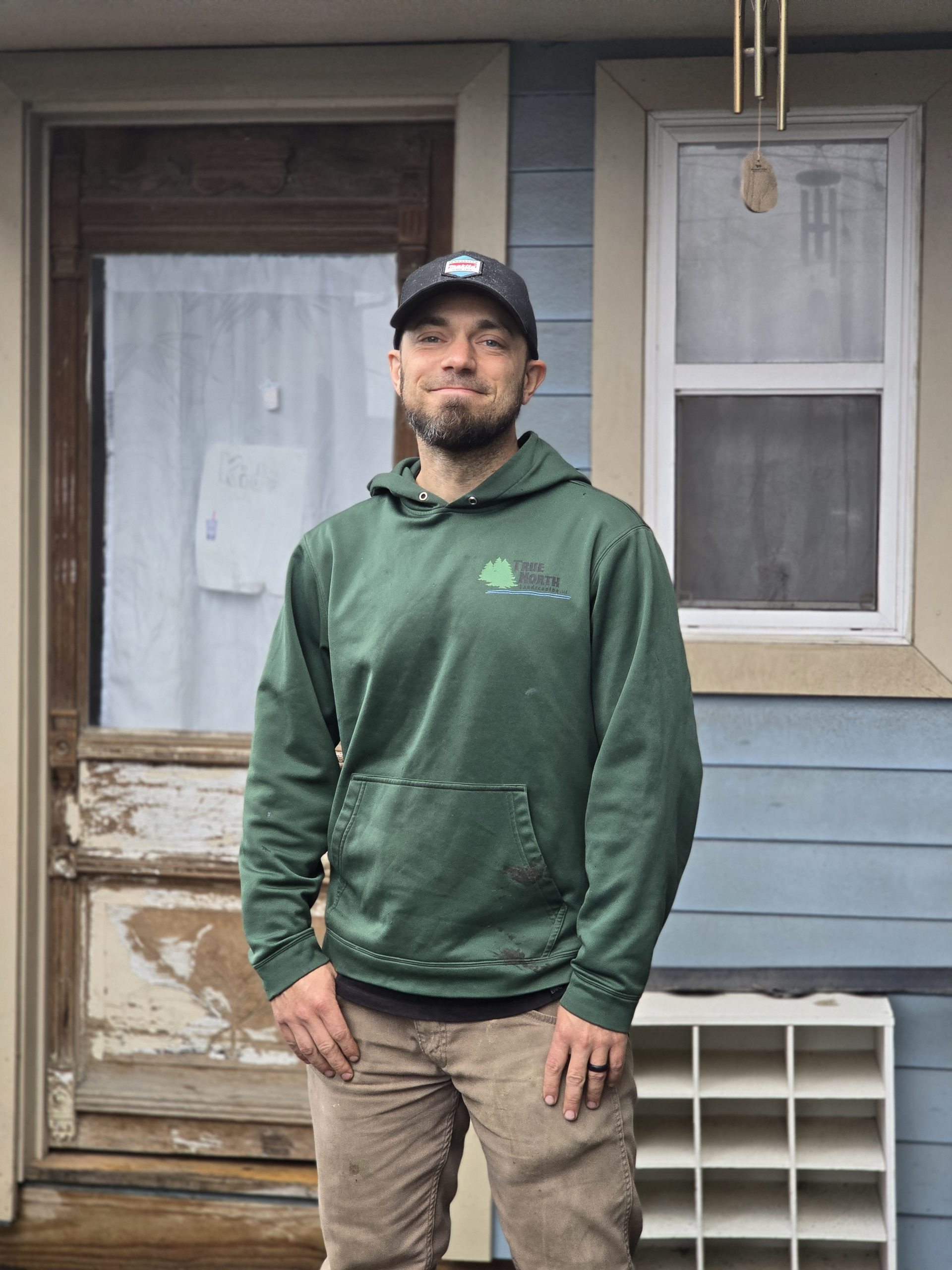 Portrait of a middle-aged male landscape architect with salt-and-pepper hair, wearing a green shirt and standing in front of a lush garden. The background is softly blurred, and the subject is smiling confidently, exuding expertise and approachability.