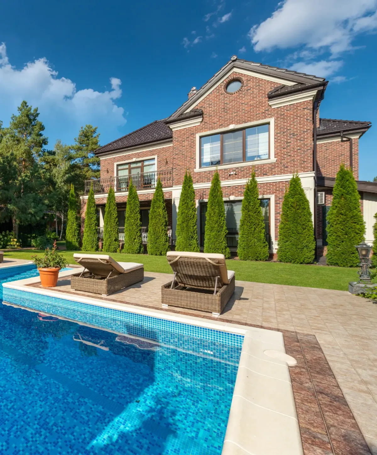 Luxury brick home with tall trees and a beautiful backyard swimming pool featuring lounge chairs on a sunny day