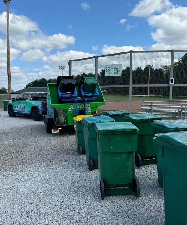 Trash can cleaning trailer servicing multiple bins near a ballfield with bleachers and a gravel lot under a partly cloudy sky 