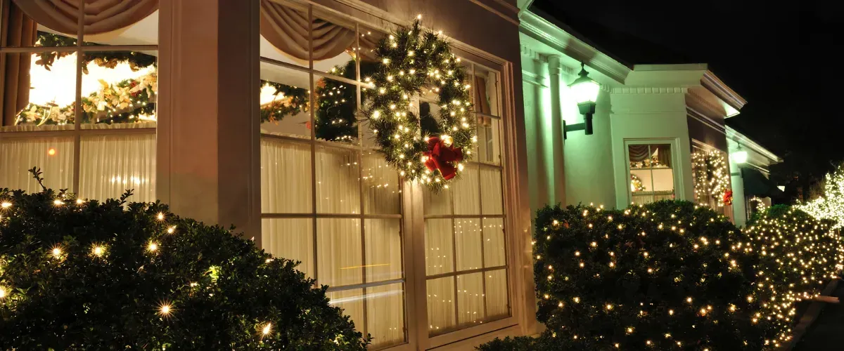 House decorated with a glowing Christmas wreath and bright holiday lights on shrubs