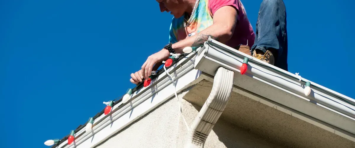 Man installing Christmas lights along the roofline of a house on a sunny day.