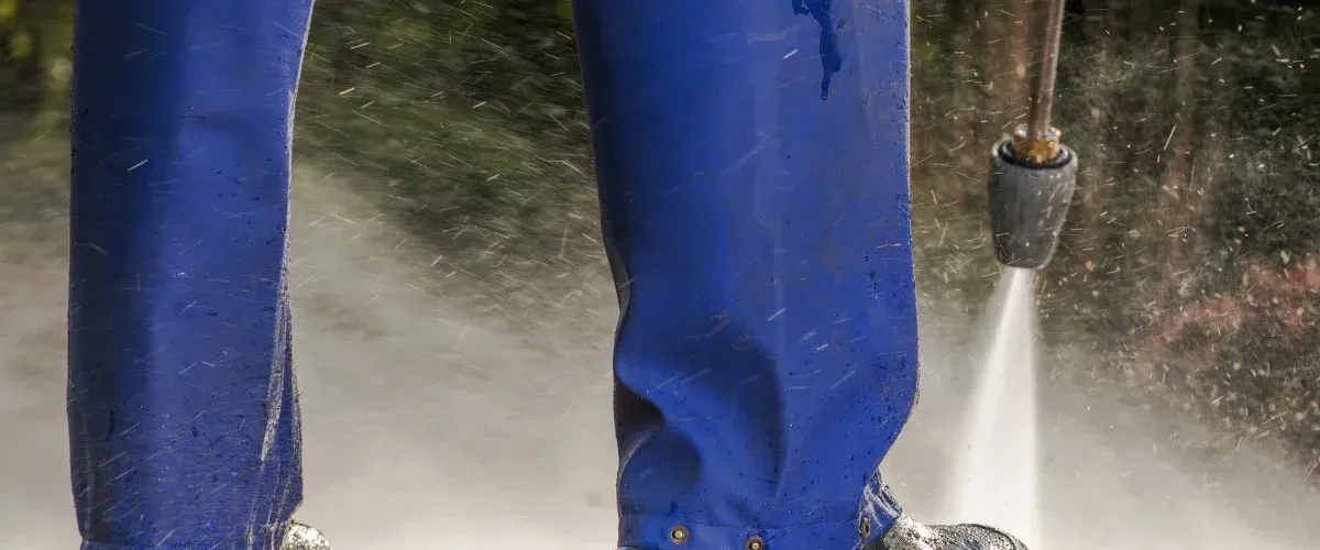 Close-up of blue protective pants and boots being splashed by water from a pressure washer nozzle.