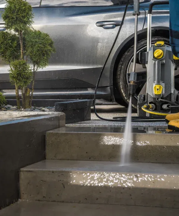 A person uses a pressure washer to clean wet concrete stairs, with a car and small bushes visible in the background.