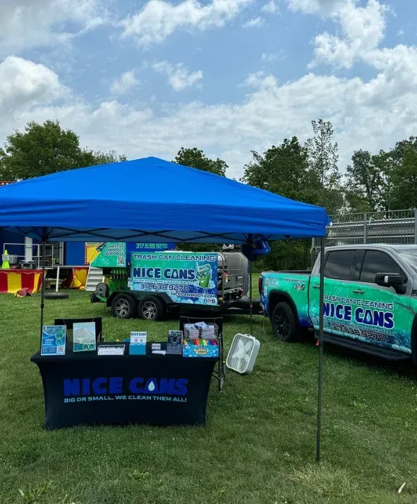 An outdoor display for Nice Cans trash can cleaning service, featuring a blue tent, table, trailer, and truck.