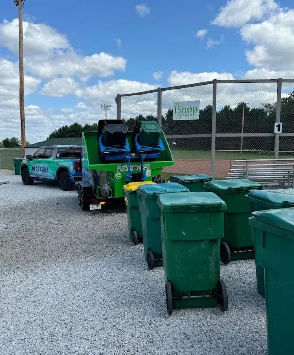 A line of large green wheeled trash cans on a gravel field with a truck and washing trailer in the background.