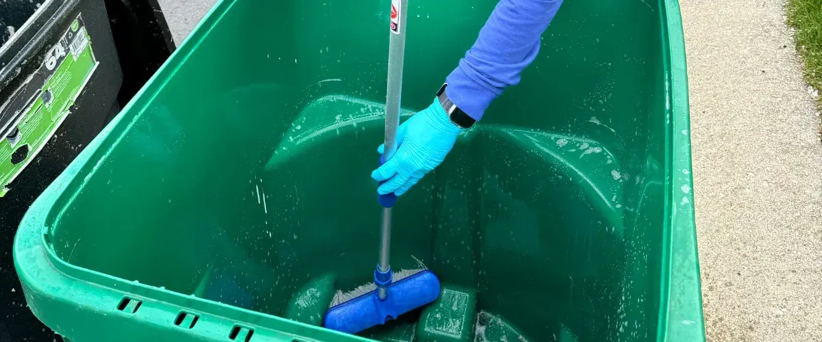 A person in blue gloves and sleeve cleaning a large green trash can with a blue scrub brush.