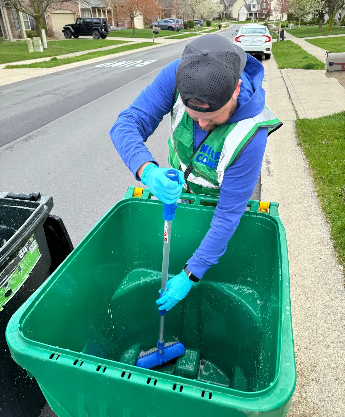 A man in blue gloves and a safety vest scrubs the inside of a large green trash bin with a long-handled brush on a residential street.