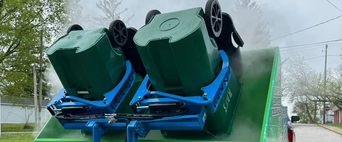 Two green wheeled trash bins are inverted for cleaning by blue hydraulic lifts on a truck, emitting mist.