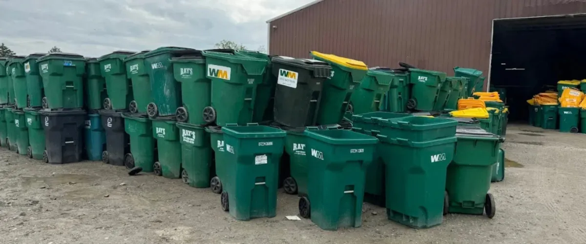 Numerous green and some black waste and recycling bins stacked outside a building under a cloudy sky.