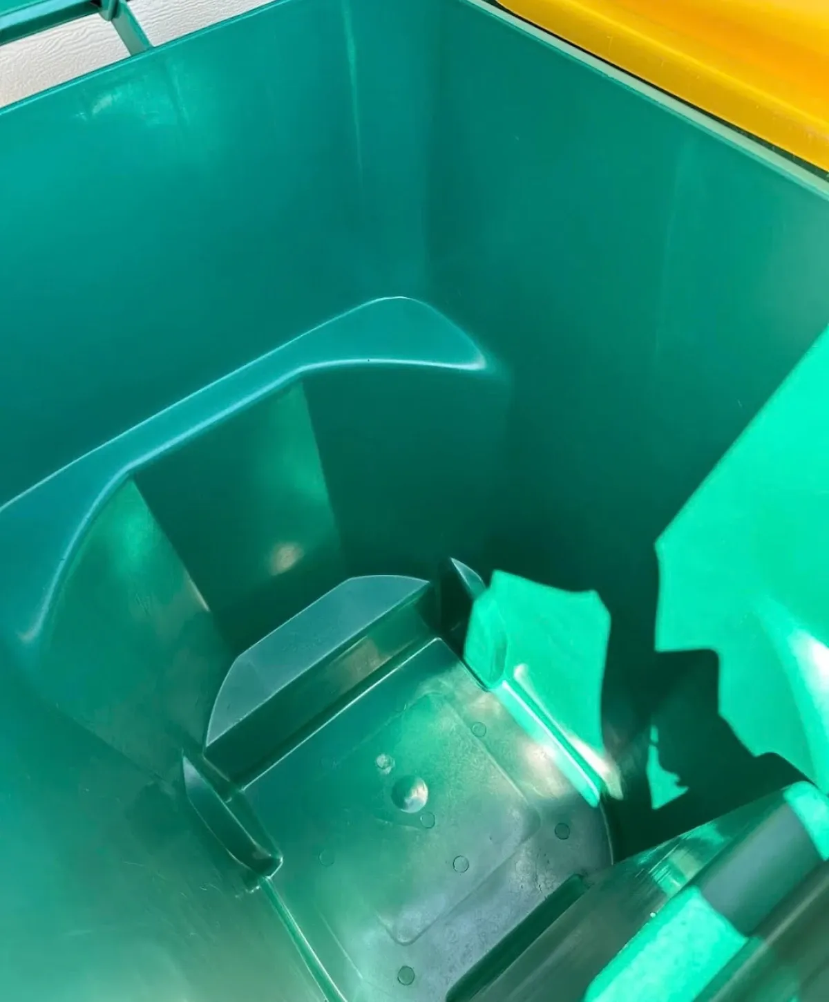 An overhead view looking down into a clean, empty, dark green plastic recycling bin with a yellow top edge.