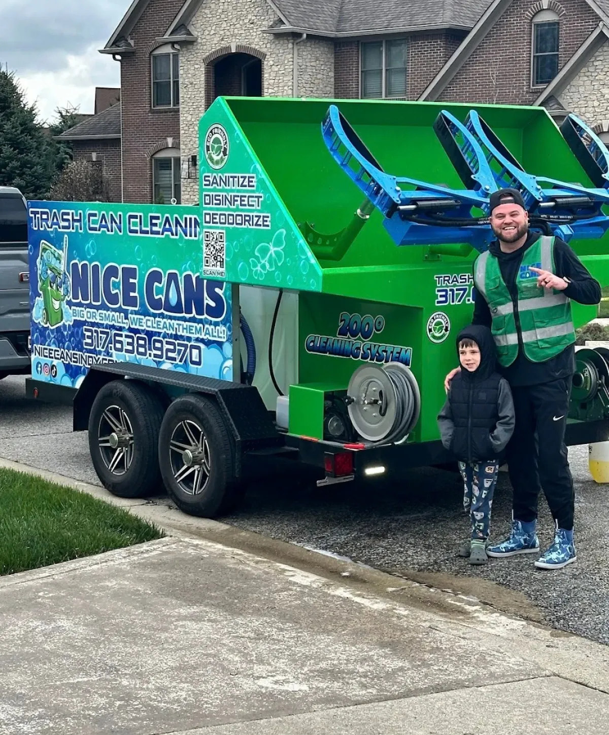 A man and young boy stand next to a green 'Nice Cans' trash can cleaning system trailer in a residential street.