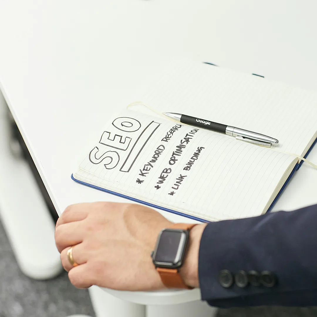 A man wearing a watch placing his hand next to a notebook that says SEO and has a list of tasks associated with search engine optimization