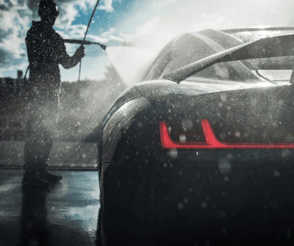 Professional car detailing in progress with a silhouette of a person power-washing a sleek black car, highlighting the vibrant red tail lights and water droplets in the air, under a bright and sunny Bend, Oregon sky.