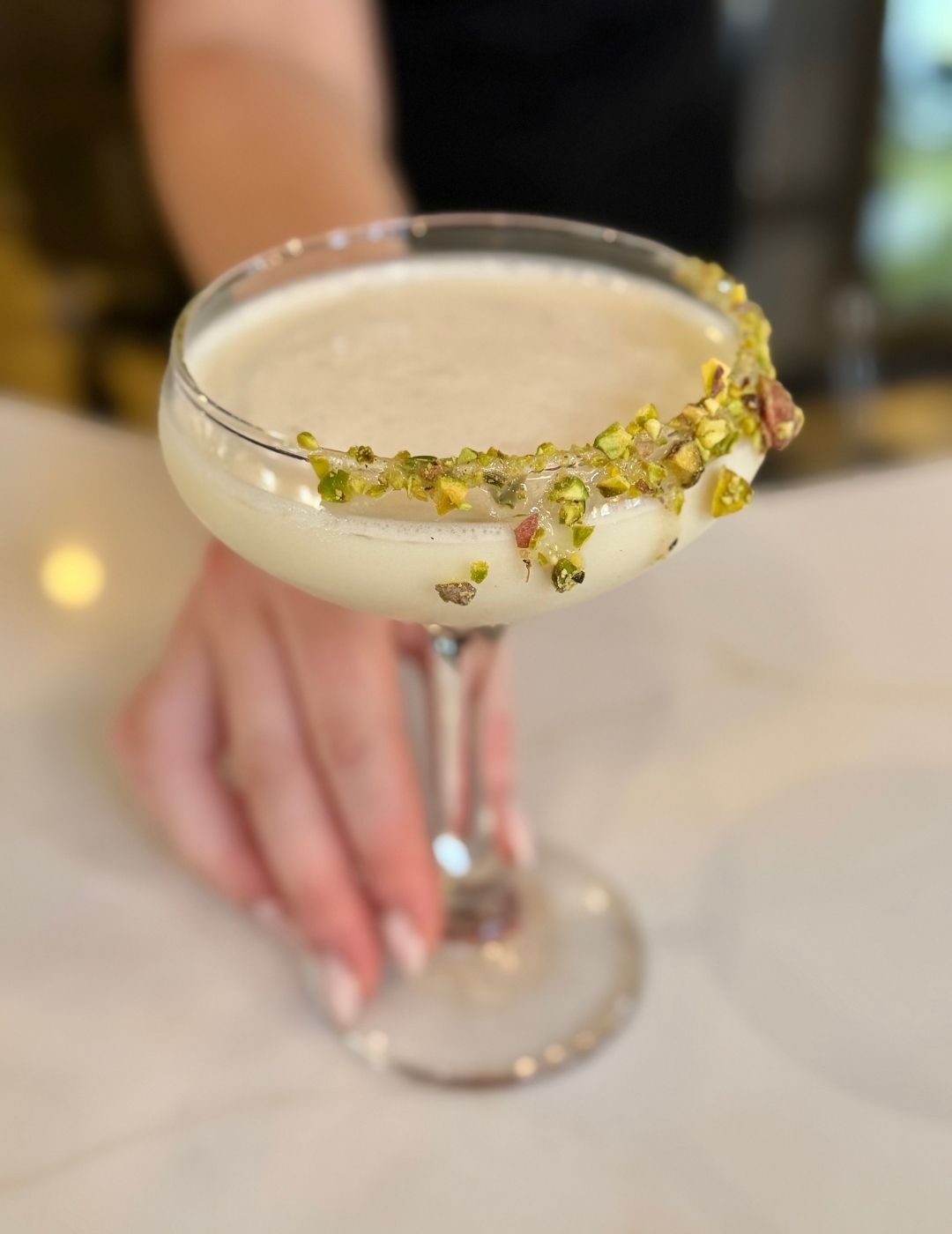 A frothy cocktail in a coupe glass held by a hand against a light background