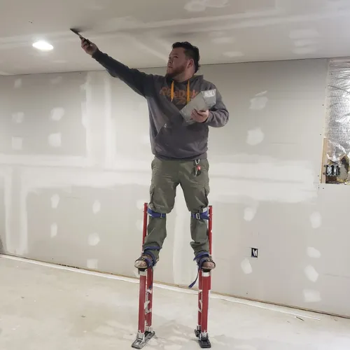 A man on stilts in a room patching a ceiling's drywall.