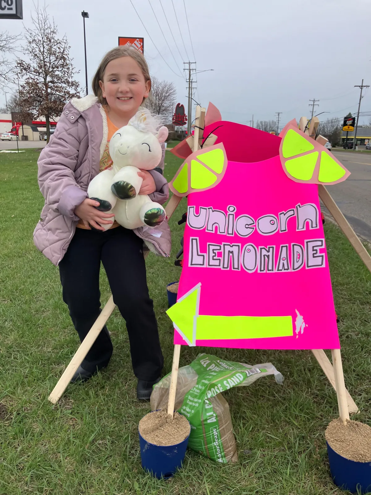 Savannah, a smiling 9-year-old entrepreneur, holding a unicorn plushie next to her bright pink 'Unicorn Lemonade' sign at her roadside lemonade stand.