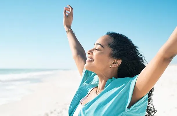 Woman standing on the beach with arms lifted smiling towards the sun.