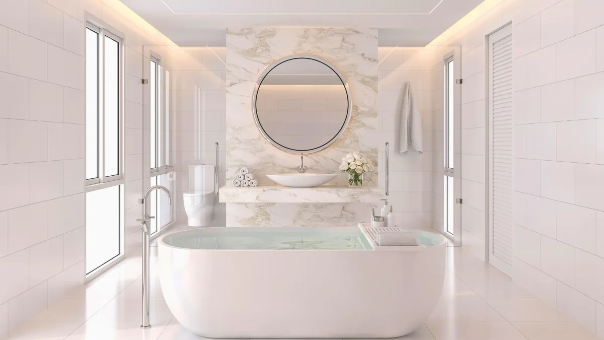 Luxurious modern bathroom with natural light featuring a freestanding white bathtub and chrome floor-mounted faucet, white vanity with a round mirror surrounded by a marble backsplash, rolled towels, and fresh flowers, all set against a backdrop of large white tiles and shutters on windows.