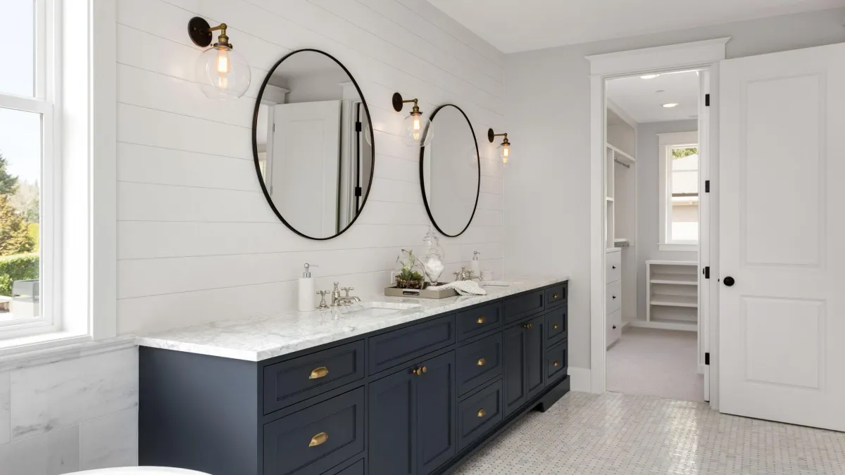 Elegant bathroom interior with his-and-her oval mirrors above a double vanity with deep blue cabinetry and brass handles, white marble countertop, traditional faucets, and decorative light fixtures, set against shiplap walls and a bright window, with a walk-in closet visible through an open door.