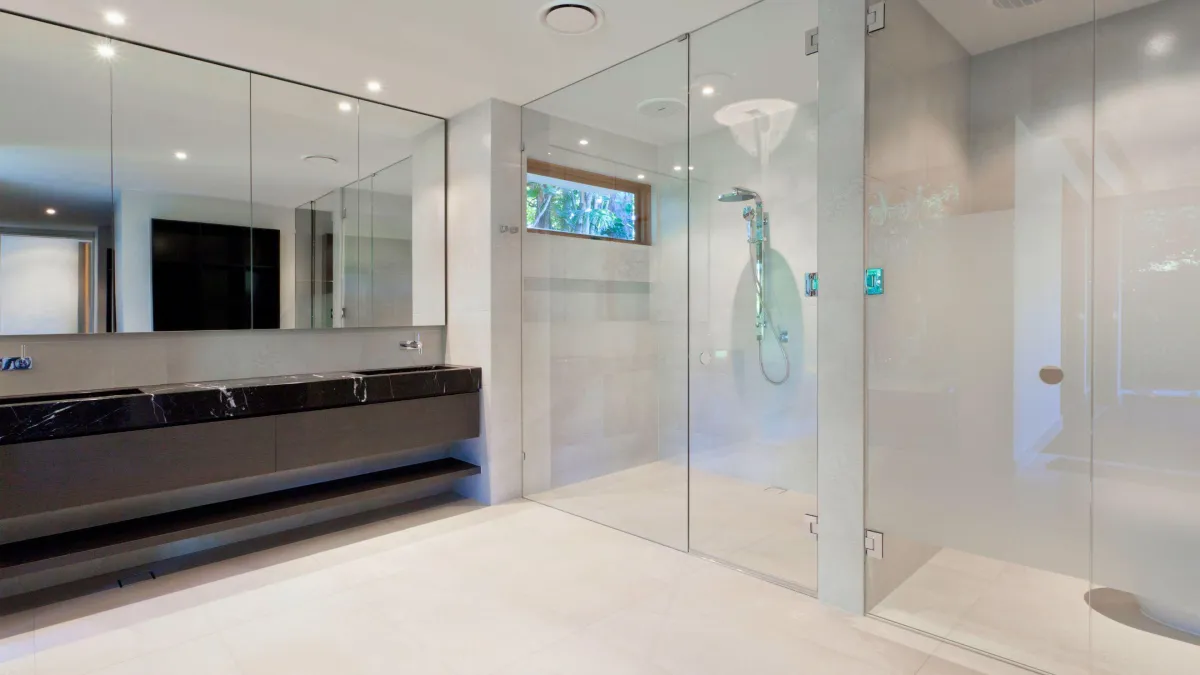 Sleek modern bathroom with a spacious glass-enclosed steam shower, his-and-hers vanity with dark marble countertop and large mirrors, reflecting a minimalist aesthetic with clean lines and a neutral color palette, complemented by natural light from a high window revealing a glimpse of greenery outside.