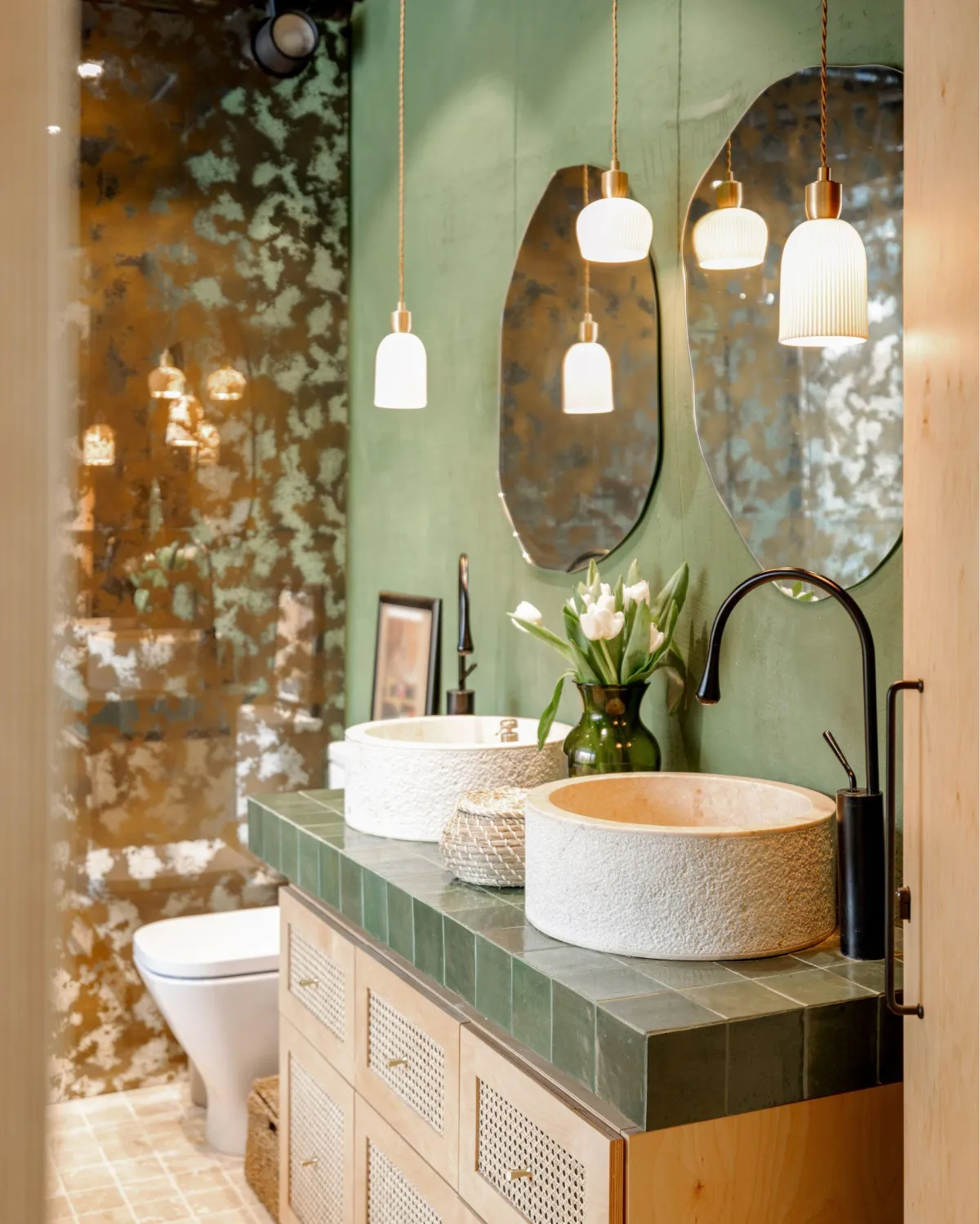 Bohemian-chic bathroom featuring dual textured stone vessel sinks atop a green tile vanity with wicker-front drawers, against an artistic oxidized copper wall creating a verdant backdrop. Pendant lights with white shades hang delicately above, and a fresh bouquet of tulips adds a touch of nature's elegance.