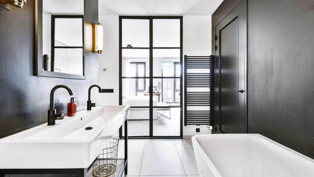 Modern minimalist bathroom with a long white his-and-hers sink vanity, featuring matte black faucets and a large mirror framed in black. A white freestanding bathtub complements the sleek design, and a tall black radiator adds a contemporary touch. A wall of black-framed glass doors leads to the adjoining bedroom, filling the space with natural light and offering a view of the urban landscape beyond.