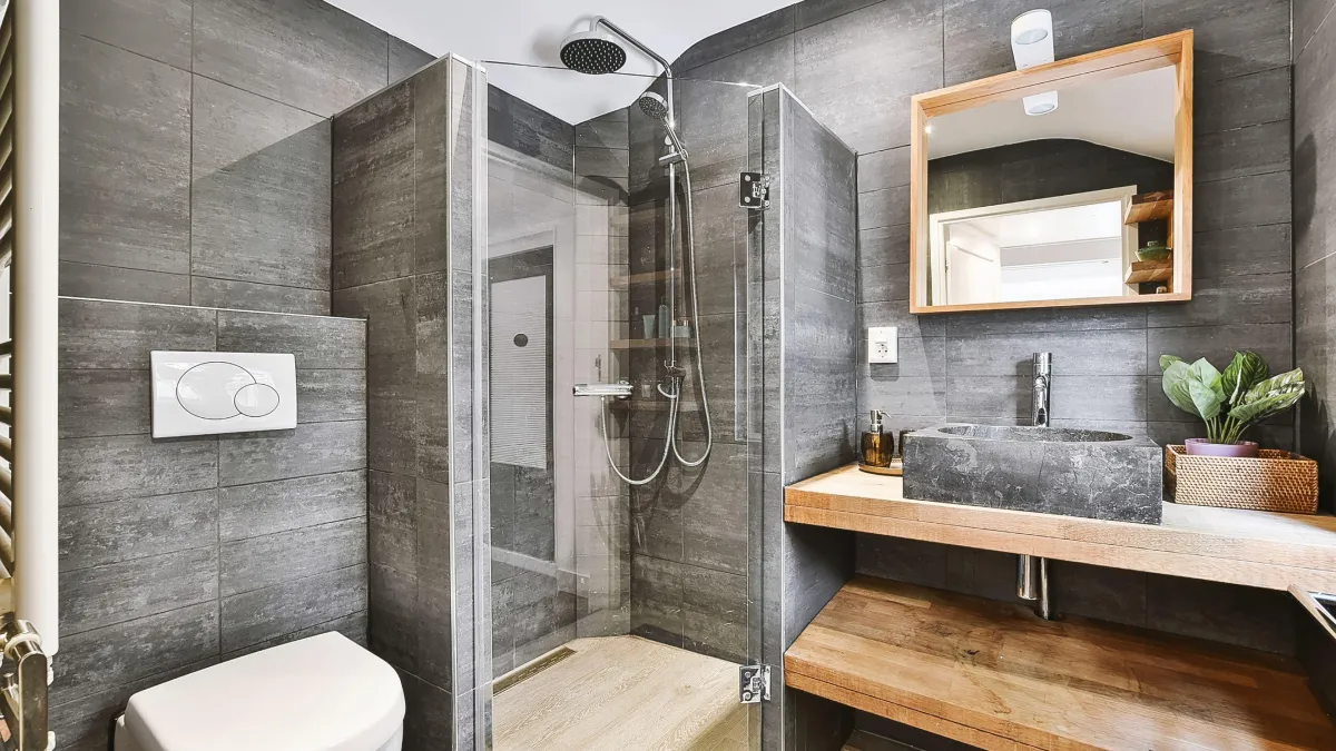 Modern urban bathroom combining raw materials, with a walk-in steam shower enclosed by transparent glass doors, surrounded by grey textured tiles. A rectangular stone sink sits on a wooden vanity against the grey tile wall, accompanied by a simple mirror with a wooden frame. Warm lighting and green plant accents add a cozy touch to the industrial-chic decor